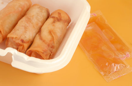 small take-out food container with spring rolls and plum sauce packets on a yellow background
