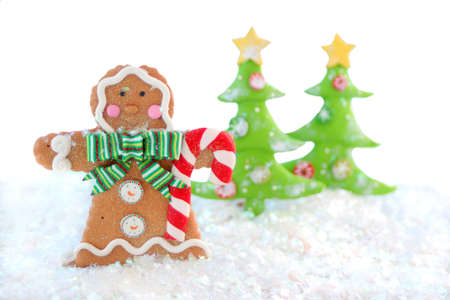 gingerbread man cookie in a winter wonderland wearing a scarf, holding a candy cane while snowing with christmas tree on a white backgroundの写真素材