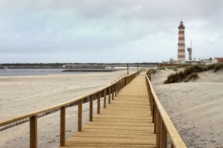 Long wooden boardwalk along the Atlantic Ocean in Praia Barra, Aveiro, Portugal with red striped lighthouse in the background on an overcast dayの写真素材