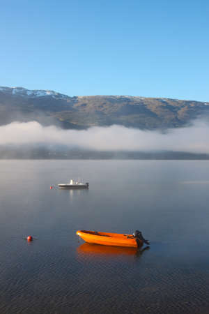 Quiet lake in rural Norway with fog patch and boats floating on a lake with mountains in the backgroundの写真素材
