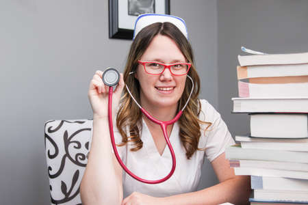 Young smiling  female nurse portrait wearing white scrubs, cap holding a stethoscope ready to check your heartbeat  with nursing textbooks on the tableの写真素材
