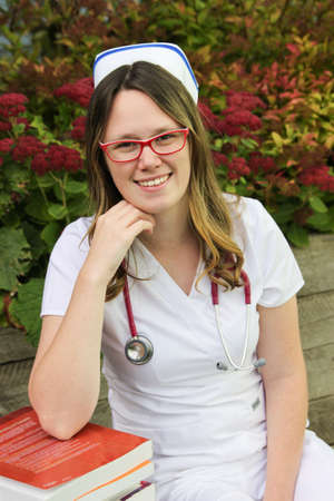 Young smiling female nurse portrait wearing white scrubs, cap and a stethoscope around her neck leaning on a stack of books outdoors during the Fallの写真素材