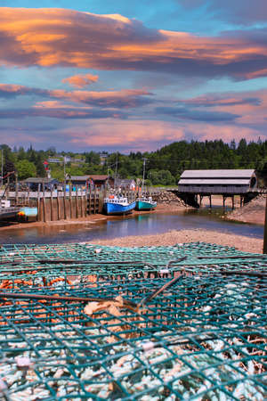Fishing wharf in St. Martins, New Brunswick, Canada on low tide with  boats in mud, a covered bridge in the background and lobster traps in front and beautiful sunset and cloudsのeditorial素材