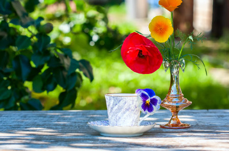 Porcelain cup of tea and beautiful spring flowers in vase on a wooden table in the garden. Summer...