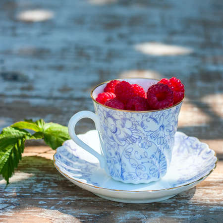 Fresh ripe raspberries and an old porcelain cup with a saucer on a wooden table in the garden. Selective focusの写真素材