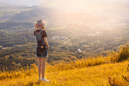 Girl with a backpack and a hat standing with her back against a background of green mountains.の写真素材