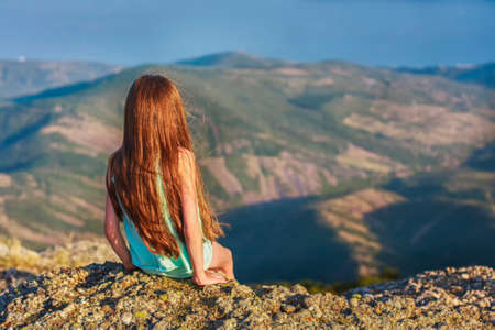 A girl with red hair sits on top of the cliff at the very edge of the abyss, admiring the scenery of the mountains, the rear view.の写真素材