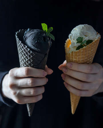 Black ice cream and white ice cream in a waffle cup in female hands. Fashionable food. Copy spaceの写真素材