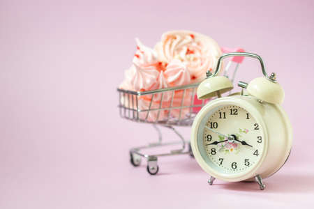 Pink mezies in a small shopping cart and an alarm clock on a pink background. Creative concept food health diet. Selective focus, copy spaceの写真素材