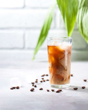 Cold brewed iced coffee in a tall glass cup and coffee beans on a gray concrete background. Pouring milk into a glass of coffee. With copy space for text.の写真素材