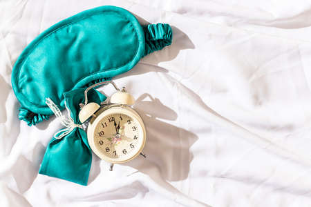Sleeping mask and alarm clock on the white silk bedding in the morning. Sweet dreams concept. Copy space, flat lay.の写真素材