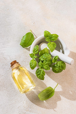 Transparent bottle of basil essential oil, fresh basil leaves in a ceramic mortar with a pestle on a light concrete background. Alternative herbal medicine and aromatherapy concept. Selective focusの写真素材