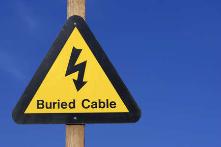 Yellow buried electrical cables warning sign against blue skyの写真素材