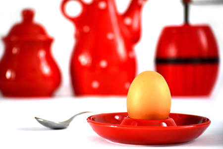 Egg on red holder near a spoon and cups and a pepper grinder in the backgroundの写真素材