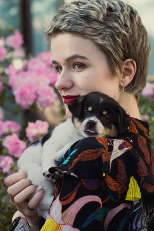 Close up blonde woman holding puppy in garden portrait picture. Closeup side view photography with flowers on background. High quality photo for ads, travel blog, magazine, articleの写真素材