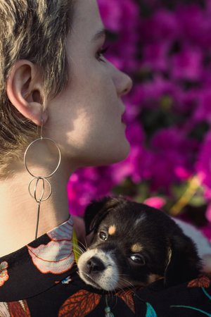 Close up puppy on shoulder of young woman in garden portrait picture. Closeup rear view photography with flowers on background. High quality photo for ads, travel blog, magazine, articleの写真素材