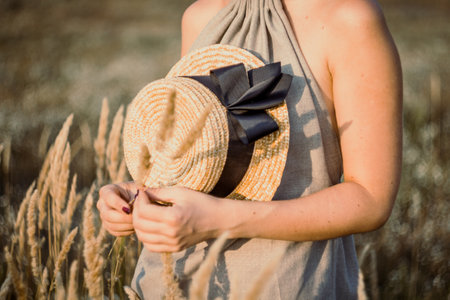 Close up pretty female hands holding straw hat with ribbon concept photo. Front view photography with grainfield on background. High quality picture for wallpaper, travel blog, magazine, articleの写真素材