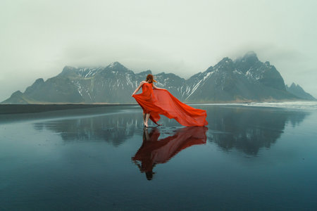Woman in waved red dress on Reynisfjara beach scenic photography. Picture of person with mountains on background. High quality wallpaper. Photo concept for ads, travel blog, magazine, articleの写真素材