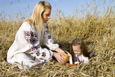 the girl pours milk baby sitting in a wheat field in national costumesの写真素材