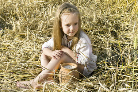 child sitting on a the field of wheat ears in national dress with a milk's pitcherの写真素材