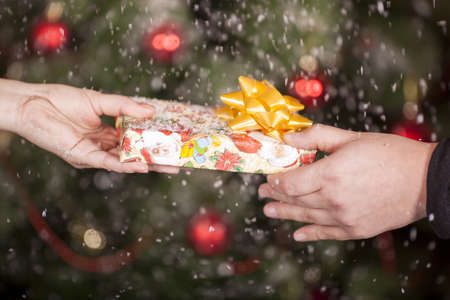 Gift delivery between a man and a woman under snowfall in front of Cristmas treeの写真素材