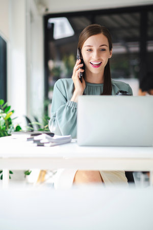 Vertical image of young confident businesswoman sitting at the office table working on laptop computer and smart phone mobile.の写真素材