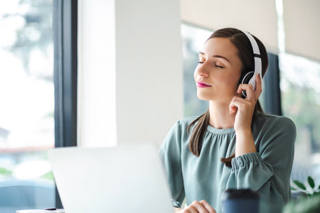 Relaxed young businesswoman in headphones enjoying music podcast using laptop app, smiling female millennial student or office worker wearing headset listening to new favorite audio tracks at work place.の写真素材
