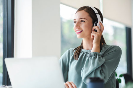 Relaxed young businesswoman in headphones enjoying music podcast using laptop app, smiling female millennial student or office worker wearing headset listening to new favorite audio tracks at work place.の写真素材