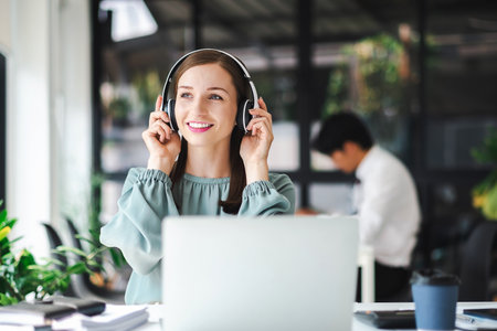 Relaxed young businesswoman in headphones enjoying music podcast using laptop app, smiling female millennial student or office worker wearing headset listening to new favorite audio tracks at work place.の写真素材