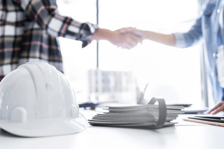 Image of engineer helmet over blur background of engineer dealings or shaking hand for architectural project. working with engineering tools on workplace.の写真素材