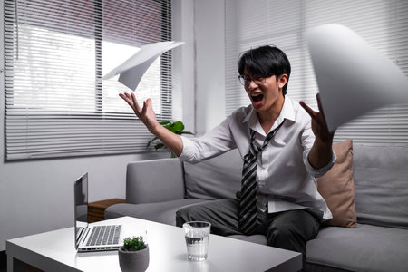 Angered, furious, crazy and mad millennial Asian businessman or man office worker screaming, shouting and throwing up papers at him office desk. Depression, overworked, failure.の写真素材