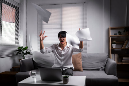 Angered, furious, crazy and mad millennial Asian businessman or man office worker screaming, shouting and throwing up papers at him office desk. Depression, overworked, failure.の写真素材