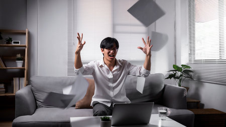 Angered, furious, crazy and mad millennial Asian businessman or man office worker screaming, shouting and throwing up papers at him office desk. Depression, overworked, failure.の写真素材