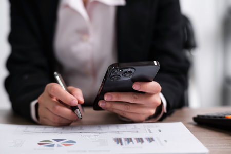 Young asian confident businesswoman sitting at the office table working on smart phone mobile and...
