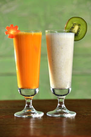 Two smoothie cocktails (kiwi fruit and vegetable carrot) in tall glasses on green background set on wooden tableの写真素材