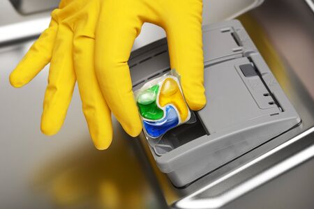 Close-up of hand in yellow rubber glove putting detergent tablet into dishwaterの写真素材