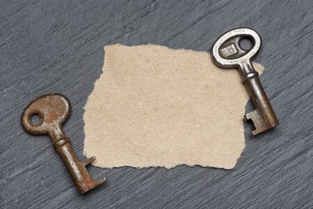 Two old rusty keys and piece of brown paper on black slate backgroundの写真素材