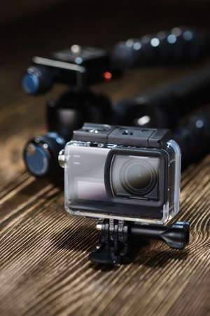 Black small action camera in waterproof case and tripod on brown wooden tabletop.