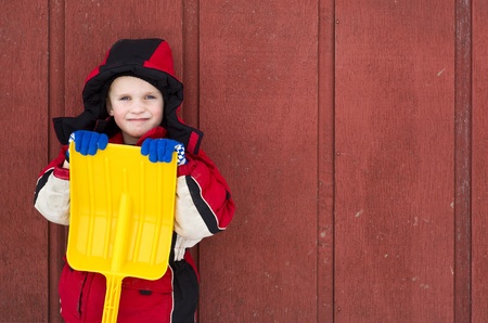 A young child, dressed in winter clothes, holds his plastic toy shovel while leaning against a red barn wall.の写真素材