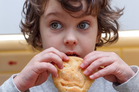 Five years old curly child eats cookies with appetite in the kitchen
