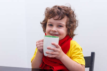 Little curly boy drinking from cup of hot drink tea in cold weather, sitting at home in scarf in kitchen at the table.の写真素材