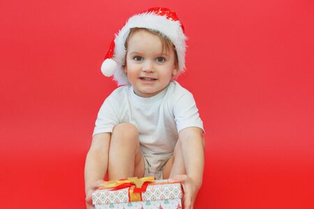 Portrait of smiling joyful child boy in Santa red hat holding Christmas gift in hand over the red backgroundの写真素材