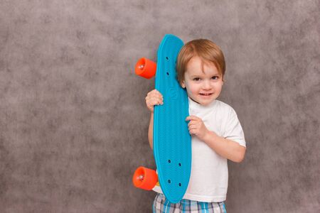 Little boy wearing a casual outfit, hold his skateboard on his shoulder, smiling, standing on a gray...