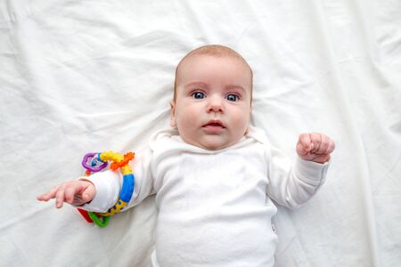 the baby smiles and lies in a white sweater on a white sheet, arms outstretched with toy. Baby with beautiful blue eyes. concept of comfort and cleanlinessの写真素材