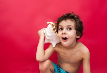 Little boy holding a big shell isolated on red background. concept of summer vacation, mobile phone,...