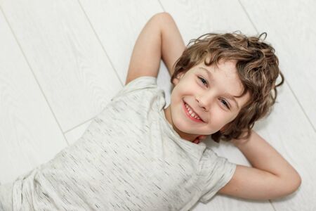 Curly boy lying on white floor at home and smiling. The concept of underfloor heating, quality laminateの写真素材