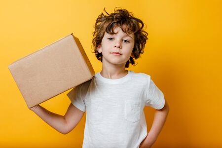 Curly Boy holds a cardboard box on his shoulder on yellow background. Concept of Food delivery, shopping online service.の写真素材