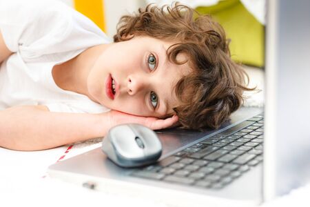 Curly boy is engaged at computer. Tired child lies covering his face with his hands Concept of...