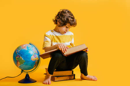 curly boy in glasses and stripes tshirt and sits on a stack of books, near to the globus on yellow backgroundm space for text.の写真素材