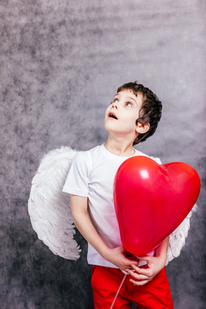 boy with wearing angel wings hid behind a red heart shaped ballの写真素材